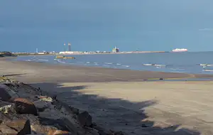Sandstrand an der Küste mit Hafen im Hintergrund, Schiffe im Wasser und eine Mole am Horizont.