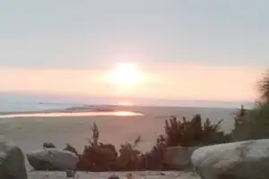 Sonnenaufgang über dem Meer mit ruhiger Bucht, im Vordergrund Felsen und Büsche am Strandweg.