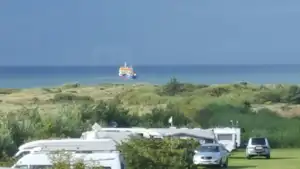 Wohnmobile und Autos auf einem Campingplatz mit Blick auf Dünen und das Meer, im Hintergrund ein Schiff.