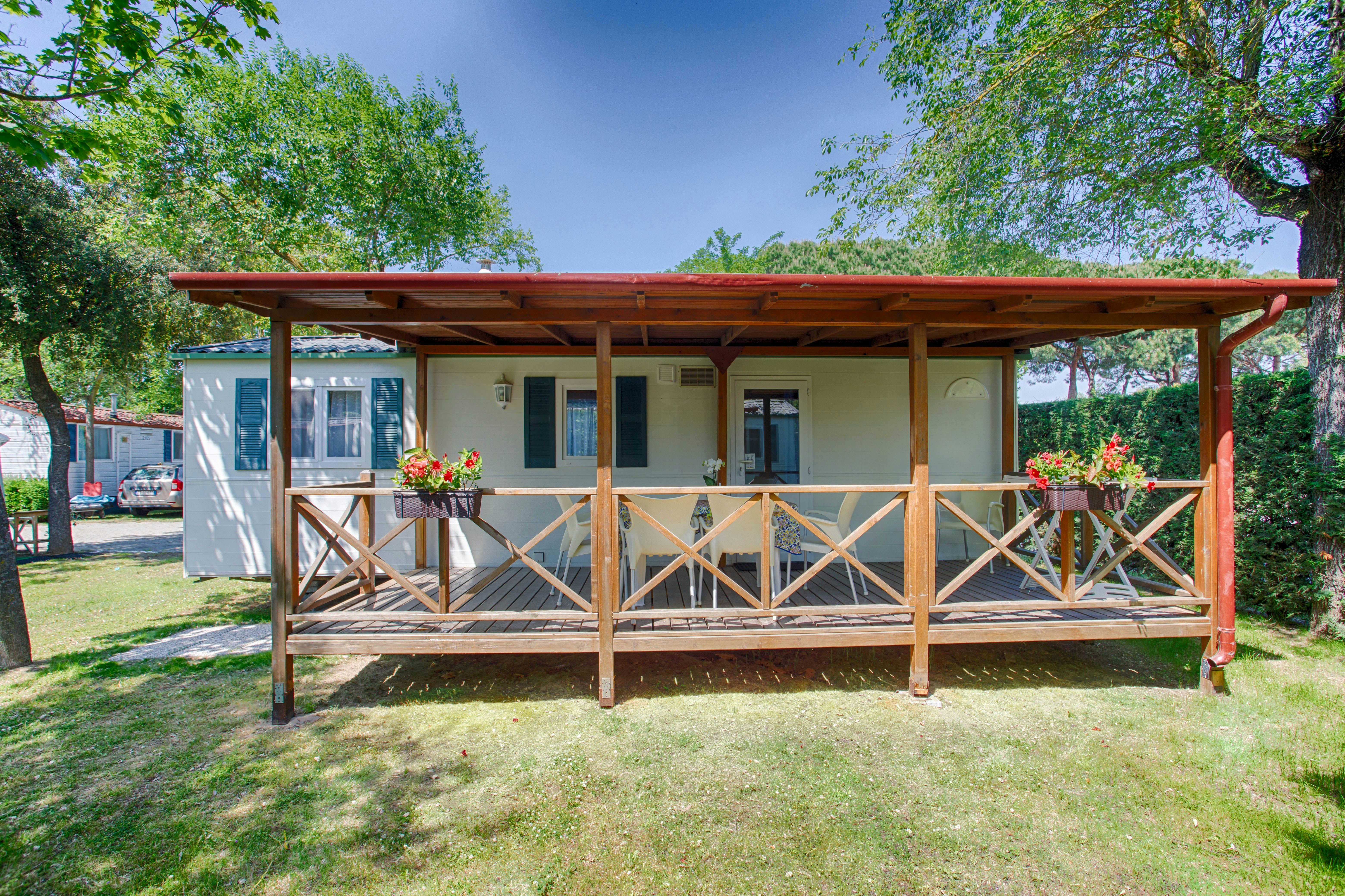 Holzveranda mit Geländer vor einem weißen Ferienhaus, Blumen im Vordergrund und Rasenfläche im grünen Garten.
