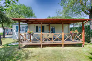 Holzveranda mit Geländer vor einem weißen Ferienhaus, Blumen im Vordergrund und Rasenfläche im grünen Garten.