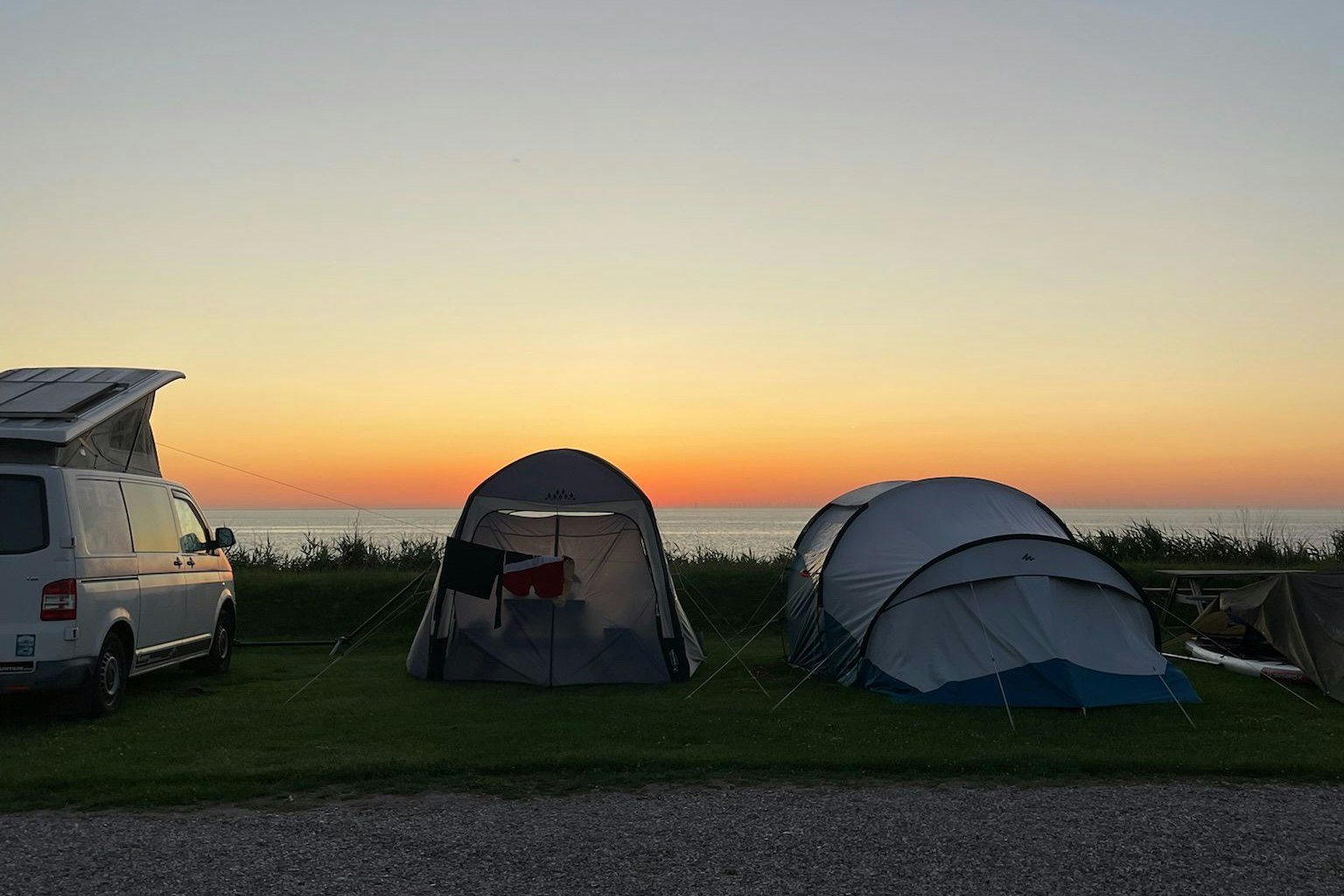 Campingplatz am Abend: Wohnmobil und Zelte auf einem Grasstellplatz, im Hintergrund der Sonnenuntergang am Horizont.