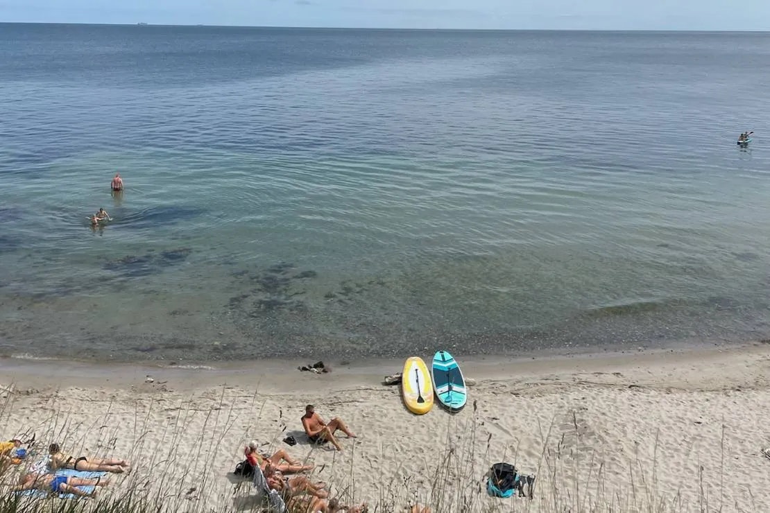 Blick auf einen Strand mit ruhigem Meer, Schwimmern im Wasser und auf dem Sand liegenden Schwimmreifen und Handtüchern.