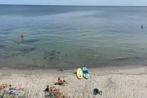 Blick auf einen Strand mit ruhigem Meer, Schwimmern im Wasser und auf dem Sand liegenden Schwimmreifen und Handtüchern.