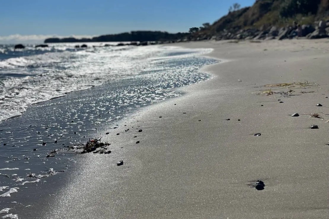 Weicher Sandstrand am Meer mit Wellen und verstreuten Steinen, im Hintergrund eine felsige Küste und bewaldete Hänge.