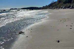 Weicher Sandstrand am Meer mit Wellen und verstreuten Steinen, im Hintergrund eine felsige Küste und bewaldete Hänge.