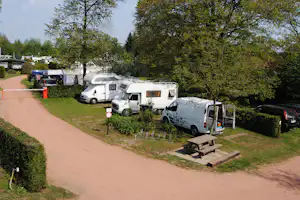 Wohnmobile und Autos auf einem Campingplatz-Stellplatz, mit Zelt und Tischbank am Weg, umgeben von Bäumen.