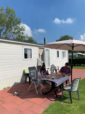 Terrasse an einem Mobilheim mit Sonnenschirm, Tisch und Stühlen vor der weißen Holzfassade, im Hintergrund Bäume und Himmel.