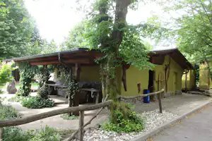 Holzunterstand und Sitzbereich am Campingplatz zwischen Bäumen, mit gepflastertem Weg, Steinmauer und gelbem Gebäude im Hintergrund.