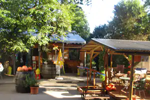 Holzhütten und überdachte Terrassen auf einem Campingplatz, mit Tischen, Stühlen, Sonnenschirm und Kübeln im Vordergrund.