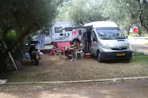 Wohnmobil und Campingbus auf dem Stellplatz, daneben Tisch und Stühle unter Olivenbäumen.