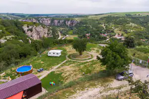 Campingplatz im Tal mit Wohnwagen auf einer Wiese, Grill und Sitzbereich, umgeben von Bäumen und Felsklippen in der Ferne.