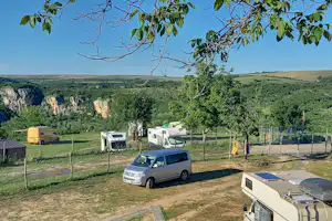 Campingplatz mit Wohnmobil und Wohnwagen auf Stellplätzen, im Hintergrund grüne Hügel und Felswände, Bäume und Wege.