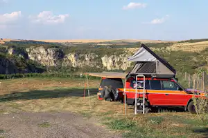 Oranger Camper mit ausklappbarem Zelt auf einem Campingplatz am Weg, im Hintergrund grüne Hügel und Felswand.