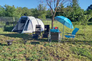 Campingzelt mit Vorzelt und Stühlen auf einer Wiese, daneben ein Sonnenschirm und ein Grill am Stellplatz.