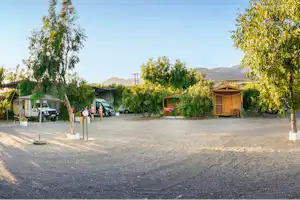 Campingplatz mit Baumreihe, Kiesweg und geparkten Transportern; rechts ein Holzschuppen mit Sitzbank, im Hintergrund Berge.