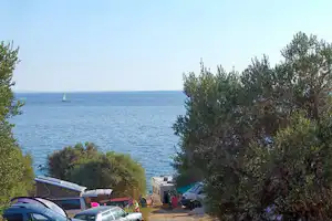 Campingplatz am Meer mit Stellplätzen, Autos und Wohnwagen, umgeben von Olivenbäumen und Blick auf das Wasser.