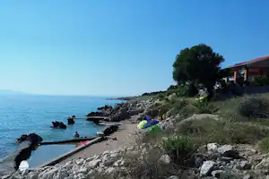 Zelt am Ufer mit Steinen und kleiner Betonrinne, Blick aufs Meer und ein Haus am Hang.