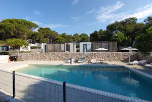 Schwimmbad mit blauem Wasser, Liegen und Sonnenschirmen vor weißen Bungalows, Steinmauer und Bäumen im Hintergrund.