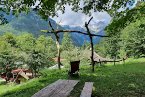 Campingwiese mit Holzbank und Weg, umgeben von Bäumen, mit Blick auf grüne Berge im Hintergrund.