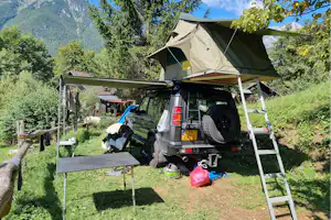 Dachzelt auf einem Campingfahrzeug mit Leiter, daneben Tisch und Stühle auf einer Wiese zwischen Bäumen und Bergen.