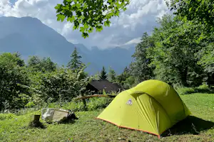 Grünes Campingzelt auf einer Wiese, daneben eine weitere Zeltplane, umgeben von Bäumen und einem Berg im Hintergrund.