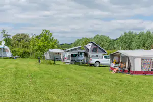 Wohnmobile und Zelte auf einem Campingplatz mit grünem Gras, Bäumen und einem Wohnwagen im Hintergrund.