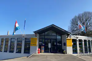 Eingang mit Rezeption und Brasserie am Campingplatz, weiße Gebäude mit Schildern, Flaggen und Sitzgelegenheiten vor der Tür.