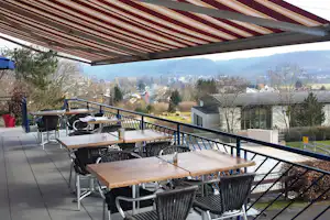 Terrasse mit Holztischen und Stühlen unter Markise, Blick ins Tal mit Häusern, Bäumen und Hügeln in der Ferne.