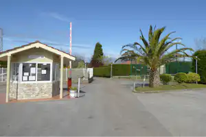 Rezeption am Campingplatz-Gebäude mit Glasfront, Schildern und Einfahrt, im Hintergrund Palmen und Hecken.