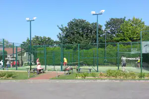 Tennisplatz mit grünen Netzen, Beleuchtungsmasten und Sitzbank am Rand, umgeben von Hecken und Bäumen.