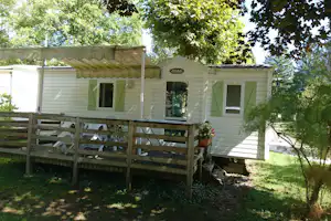 Mobilheim mit Terrasse, grünen Fensterläden und Markise, umgeben von Bäumen und Holzgeländer am Campingplatz.