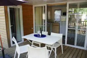 Terrasse am Holzbungalow mit weißem Tisch, Gläsern und Flasche, Blick auf Küche und große Schiebetür zum Balkon.