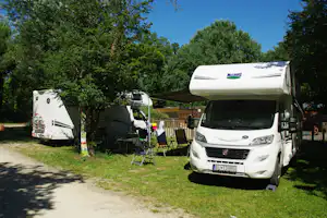 Wohnmobil mit Markise und Vorzelt auf dem Campingplatz, daneben Stühle und Tisch, umgeben von Bäumen und Rasen.
