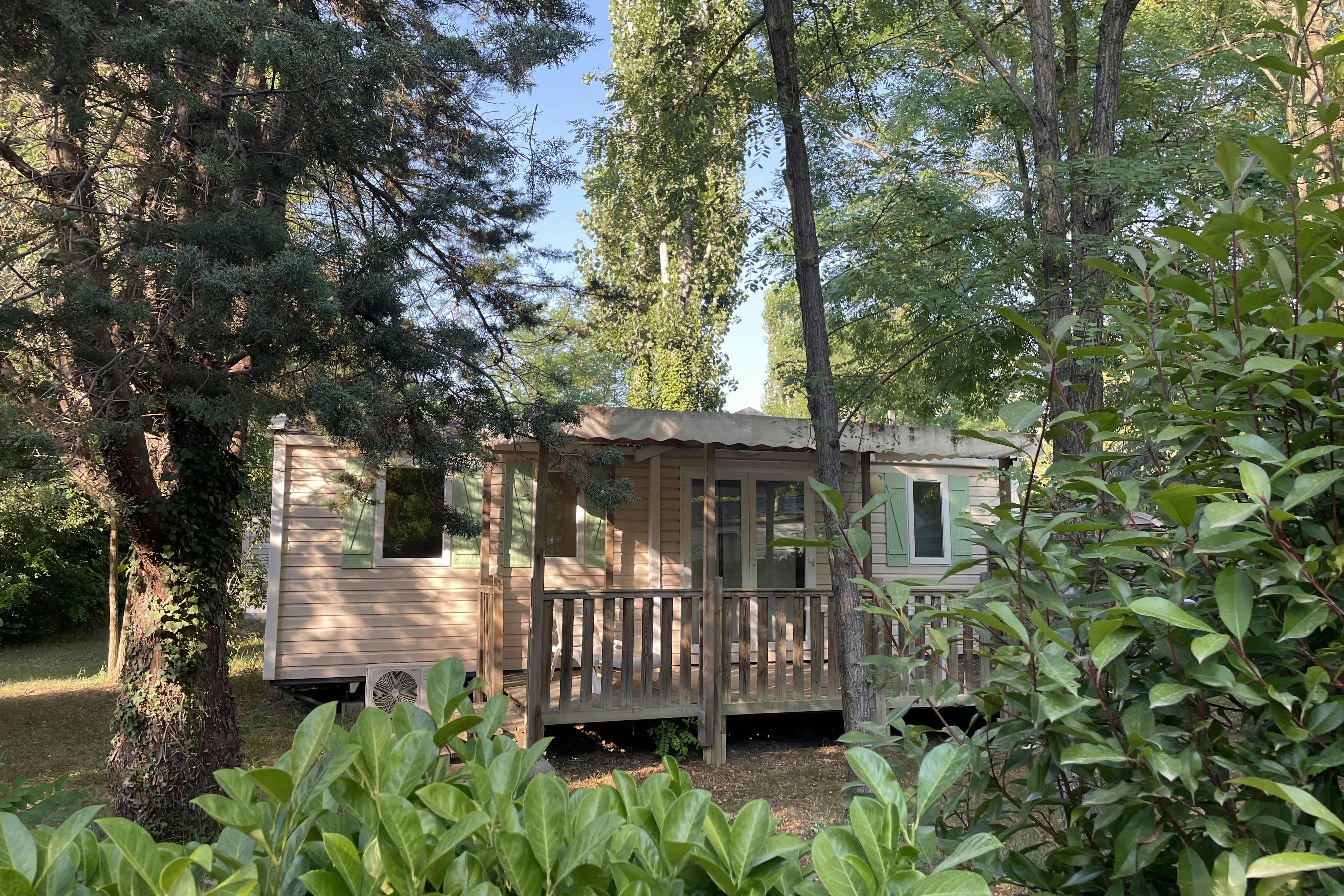 Holzhütte auf einem Campingplatz mit Veranda und Holzgeländer, umgeben von Bäumen und Sträuchern.