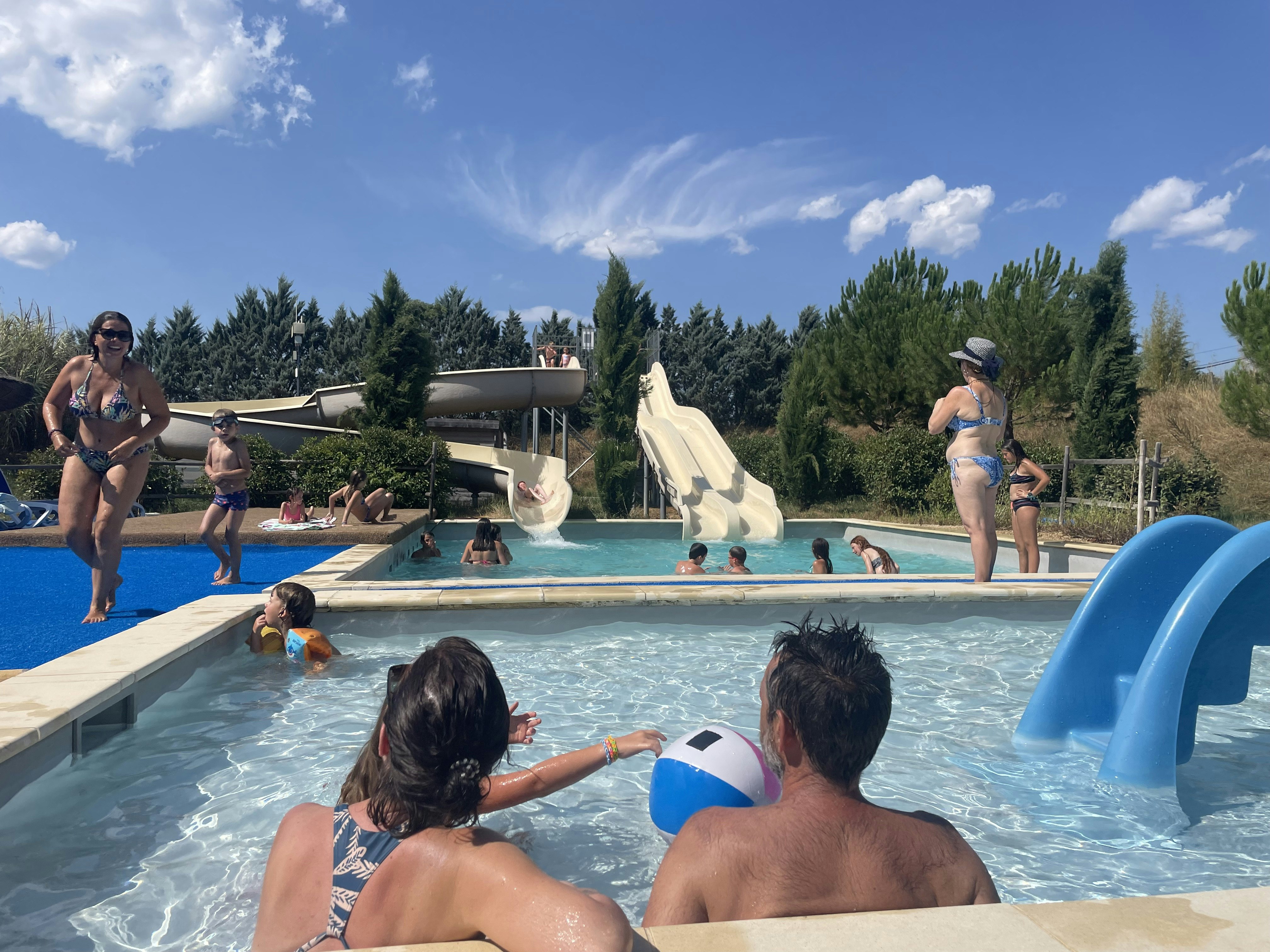 Kinder und Erwachsene im Pool eines Campingplatzes, mit Rutschen und Liegestühlen am Becken, im Hintergrund Bäume und Gebäude.