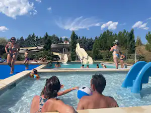 Kinder und Erwachsene im Pool eines Campingplatzes, mit Rutschen und Liegestühlen am Becken, im Hintergrund Bäume und Gebäude.