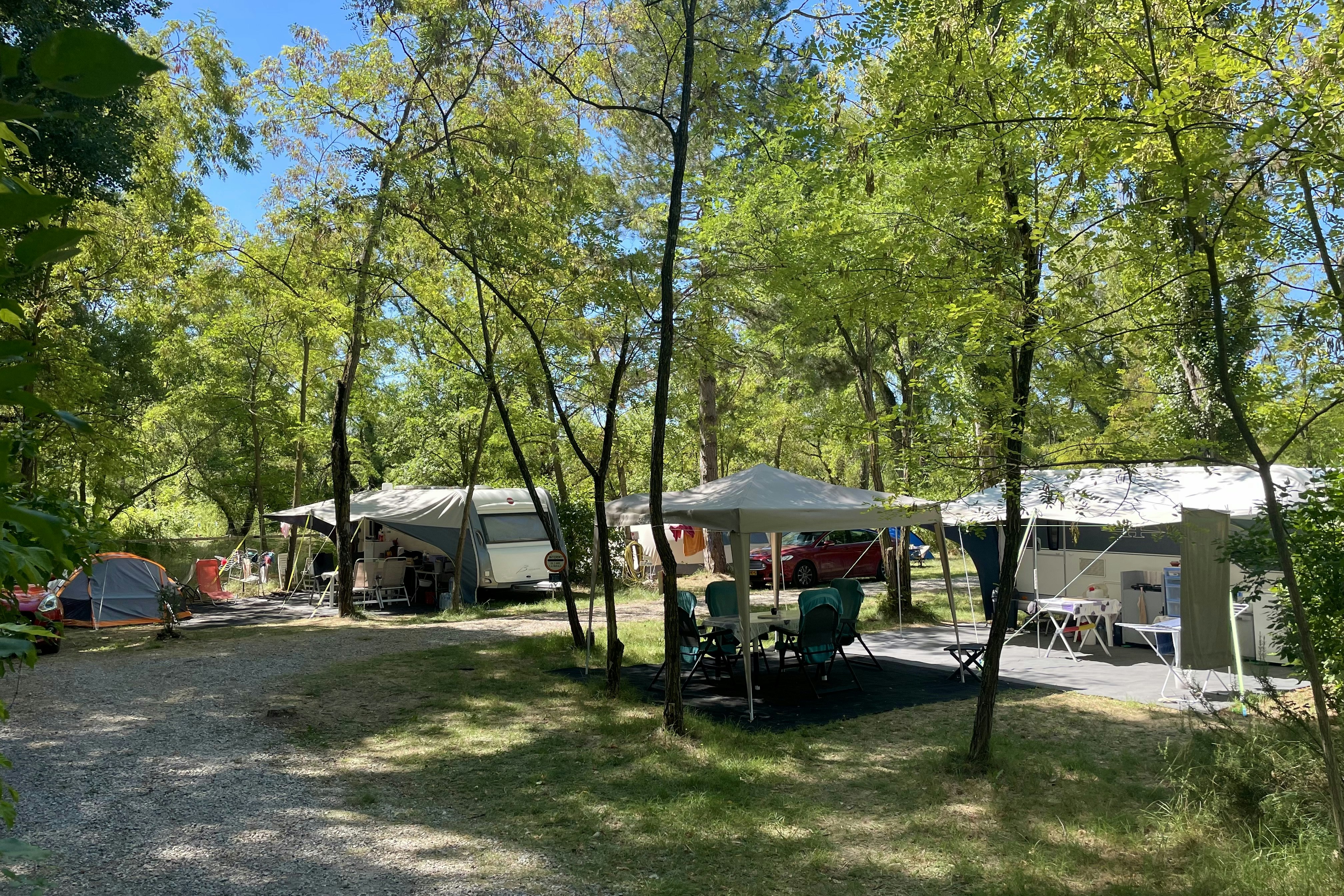 Campingplatz mit mehreren Zelten im Grünen, Sitzgruppe und Tisch unter einem Sonnenschirm zwischen Bäumen.