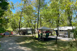 Campingplatz mit mehreren Zelten im Grünen, Sitzgruppe und Tisch unter einem Sonnenschirm zwischen Bäumen.