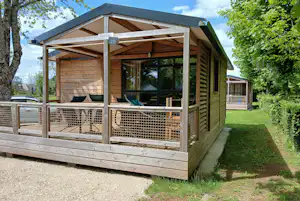 Holzbungalow mit Veranda, Sitzbank und großen Fenstern, umgeben von Bäumen und gepflegtem Rasen am Campingplatz.