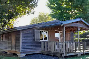 Holzhütte mit Veranda und Geländer, seitlich am Gebäude, umgeben von Bäumen und Grünfläche.