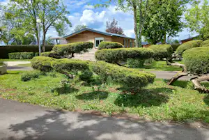Campinghaus mit Holzveranda und gepflegtem Garten mit Hecken, Bäumen und Kiesweg vor dem Gebäude.