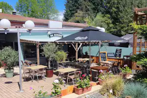 Terrasse am Campingplatz mit Tischen und Stühlen unter Sonnenschirmen vor einer Bar-Restaurant-Wand.