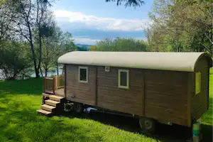 Holz-Wohnwagen mit weißem Dach, Treppe und kleiner Terrasse im Grünen, Bäume und Blick auf den See im Hintergrund.