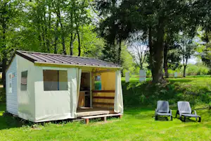 Camping-Unterkunft mit offenem Vorzelt und Holzpodest, zwei Gartenstühle auf grünem Rasen, umgeben von Bäumen.