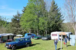 Campingplatz mit geparkten Autos, Zelt und Wohnwagen auf einer Wiese, umgeben von Bäumen und einem blauen Zelt.