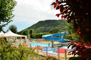 Camping-Pool mit blauen Sonnenschirmen, Liegen und Bergblick, im Hintergrund ein weißes Gebäude und Bäume im Vordergrund.