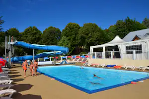 Blaues Camping-Schwimmbecken mit Wasserrutsche, Liegen und Sonnenschirmen, im Hintergrund ein weißes Gebäude und Bäume.