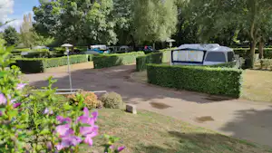 Campingplatz mit gepflastertem Weg, Hecken und einem Wohnwagen-Zeltanhänger, im Hintergrund Bäume und weitere Stellplätze.