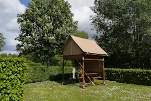 Ein Zelt unter einem Holz-Unterstand auf einer Wiese, umgeben von Bäumen und Hecken.